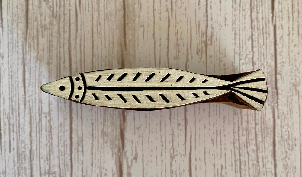 A small wooden fish-shaped object with carved geometric patterns, photographed from above on a light wood-grained surface.