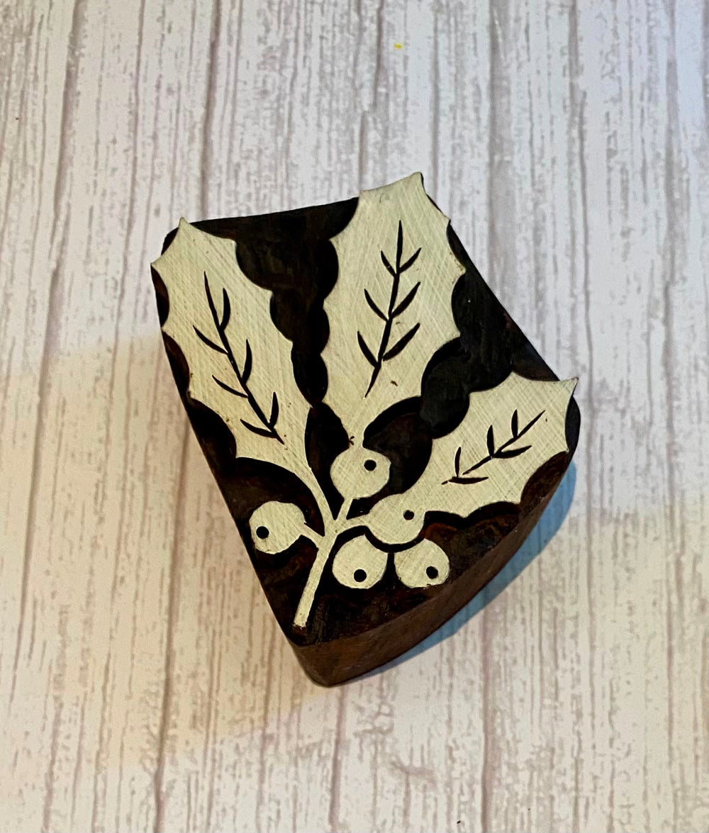 A dark brown wooden block with an inlaid cream-colored holly leaf and berry pattern, photographed on a light wood-grained surface.